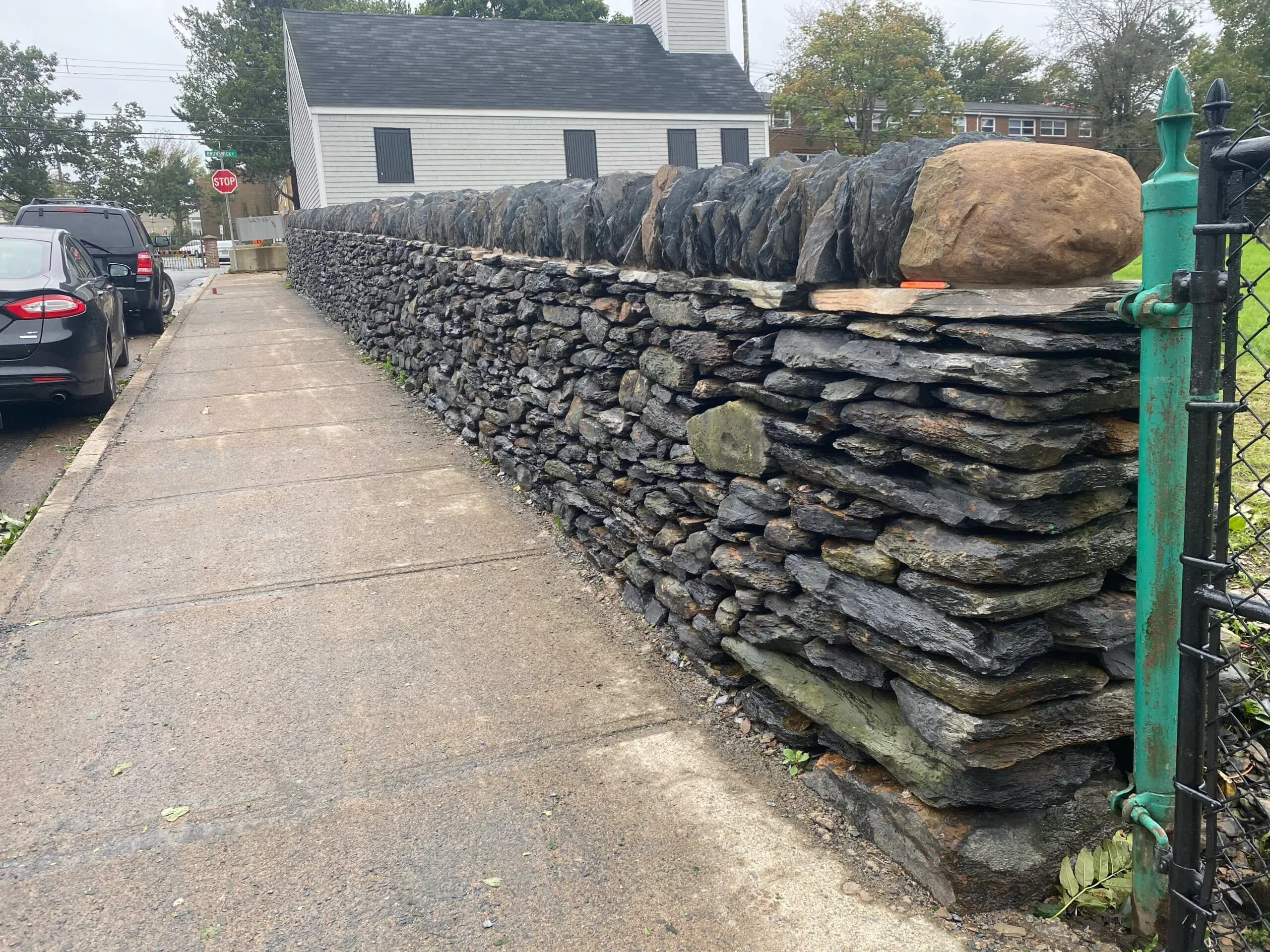 The rebuilt wall running away up the sidewalk towards the white clapboard church, its coping stones laid along the top and a green iron gate post at the near end.