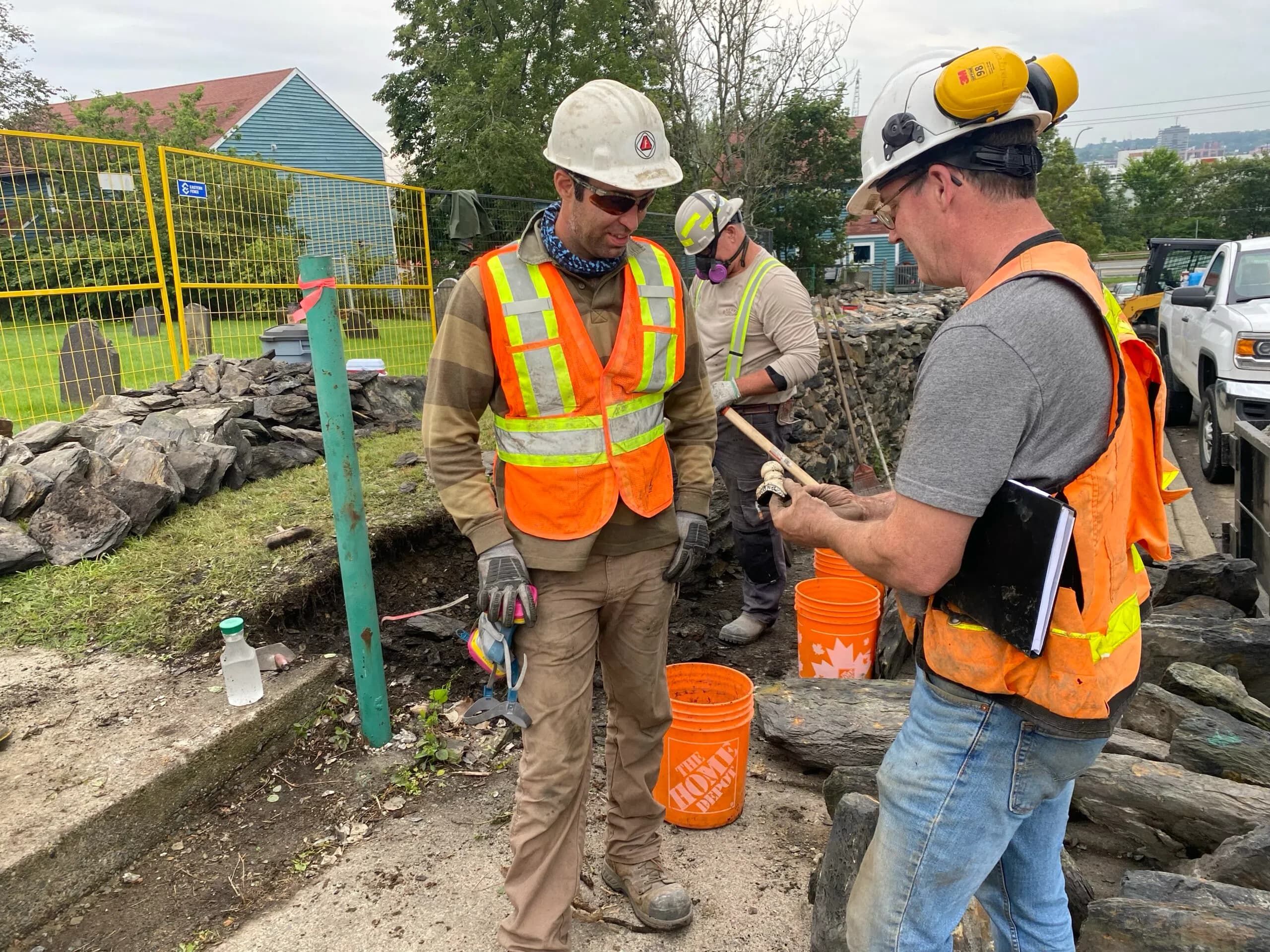 Three people in hard hats and hi-vis examining a small find held in one of their hands, the burial ground and its headstones behind the fence at the left.