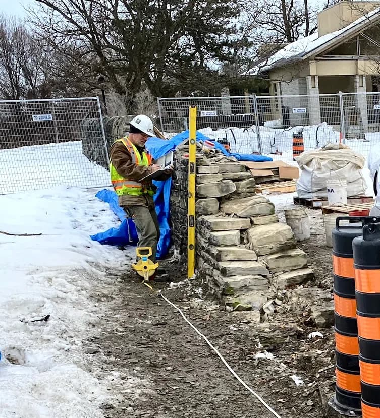 A waller in hi-vis and a hard hat writing on a clipboard at the end of the wall in winter, a levelling staff propped against the stonework and snow across the site behind.