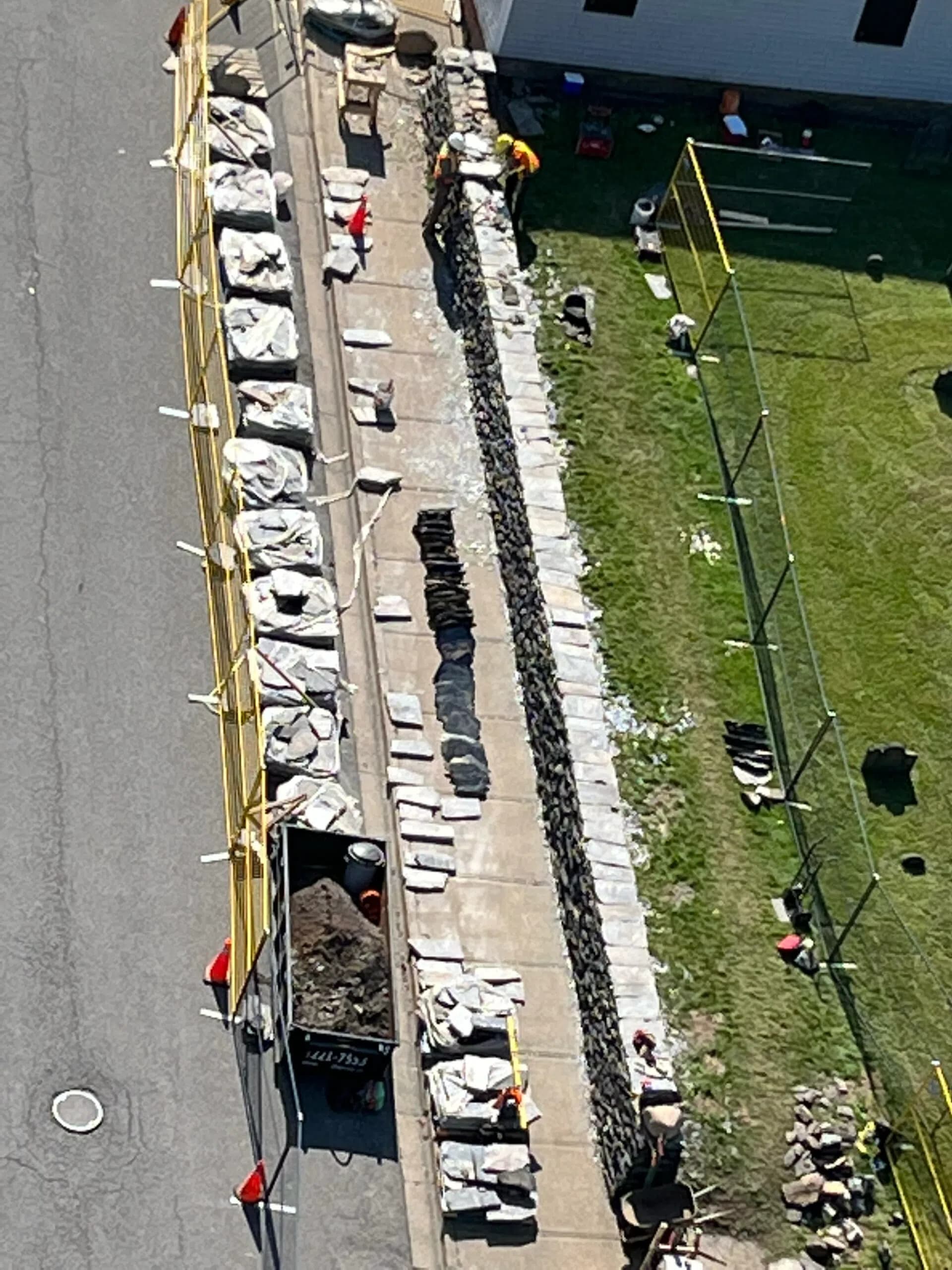The site from above: the line of the wall running the length of the sidewalk, its stone lifted out and stacked in rows along the kerb, the open foundation trench beside it and the burial ground on the far side.