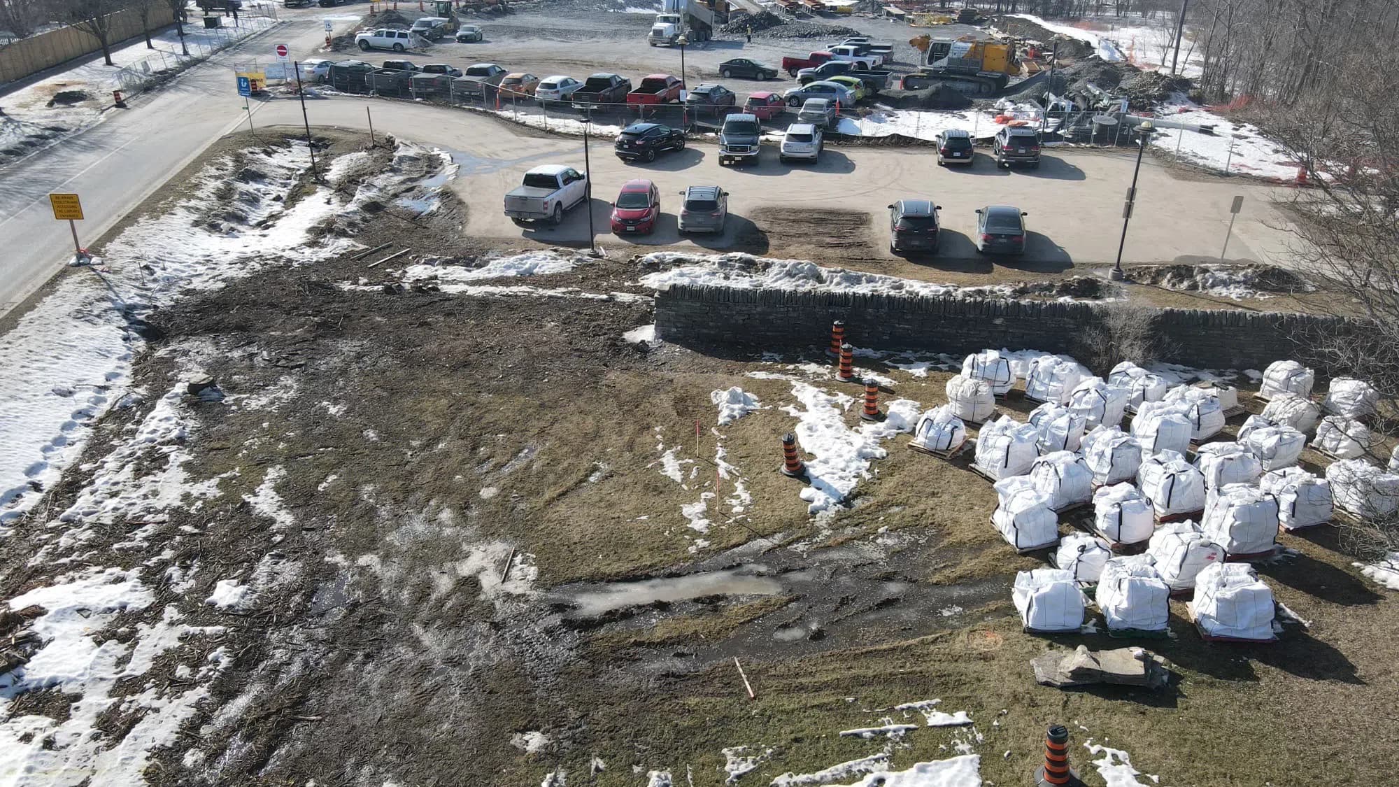 An aerial view of the site in winter: the bagged stone standing in rows of white pallet bags on the frozen ground beside the remaining length of wall, with the library car park beyond.