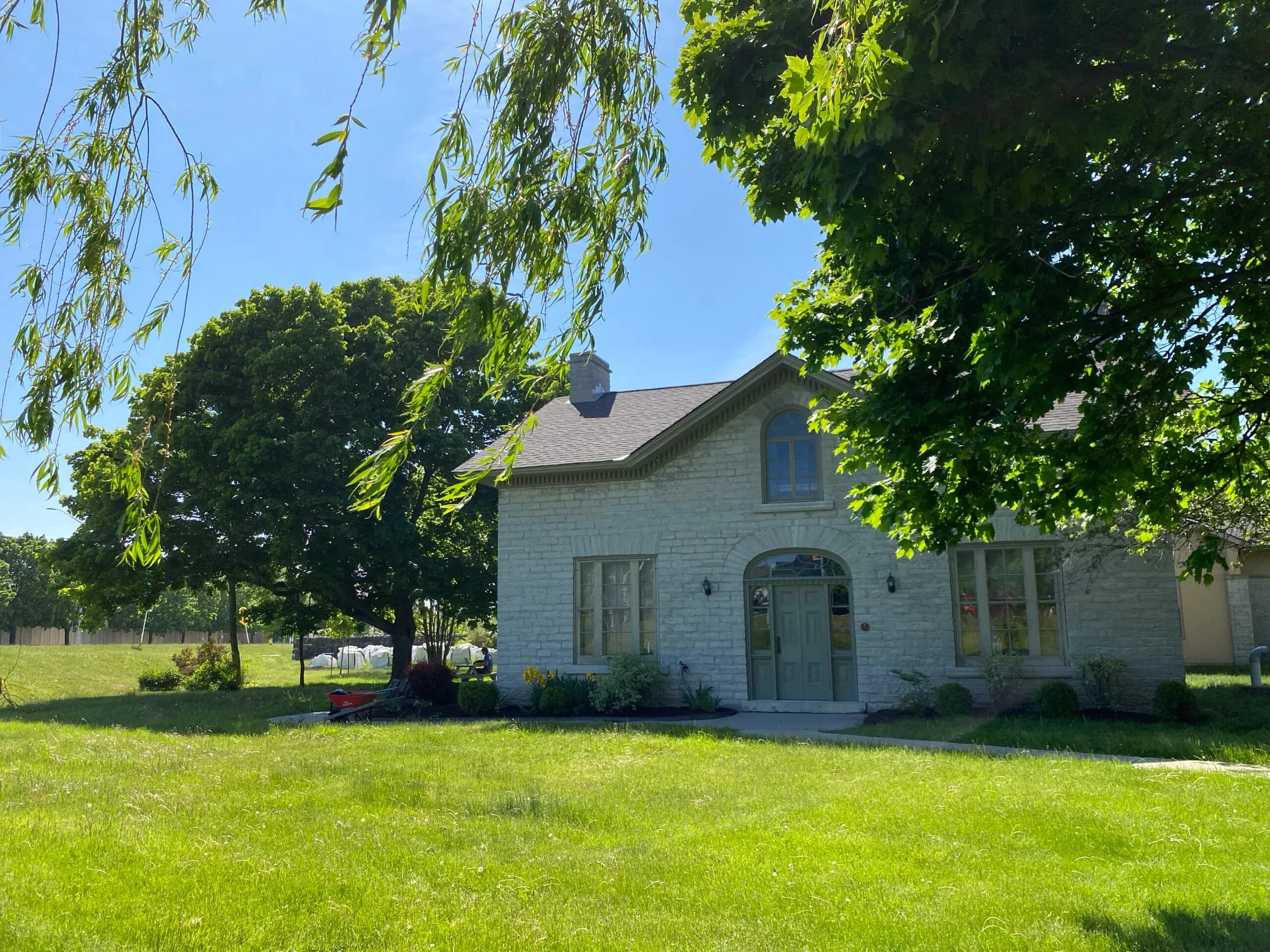 The c1860 limestone farmhouse that houses the library branch, its arched doorway and stone façade framed by mature trees on a bright summer day.