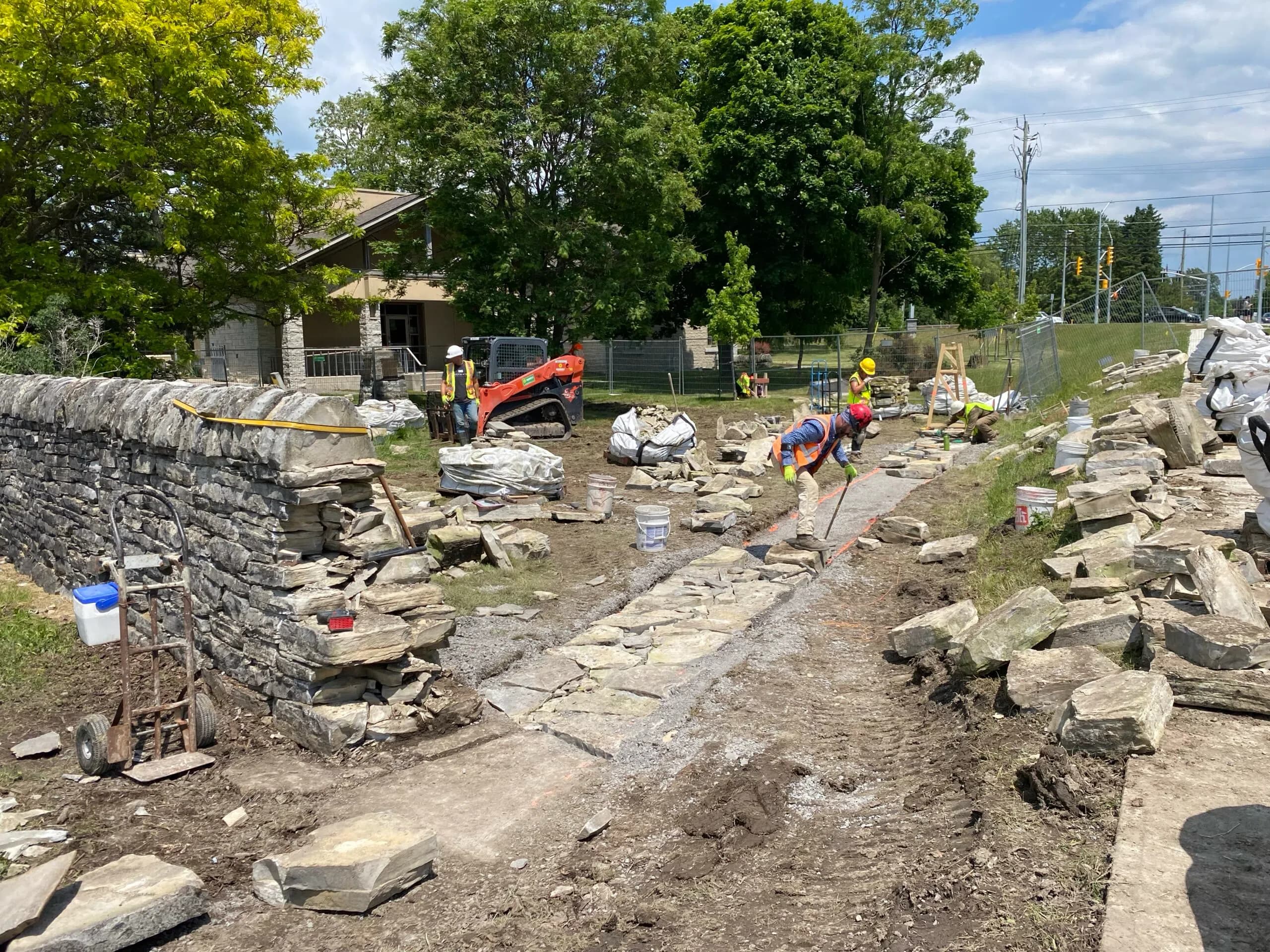 The excavated foundation trench running the length of the new wall line, the mini-excavator and crew working along it and the library visible through the trees behind.