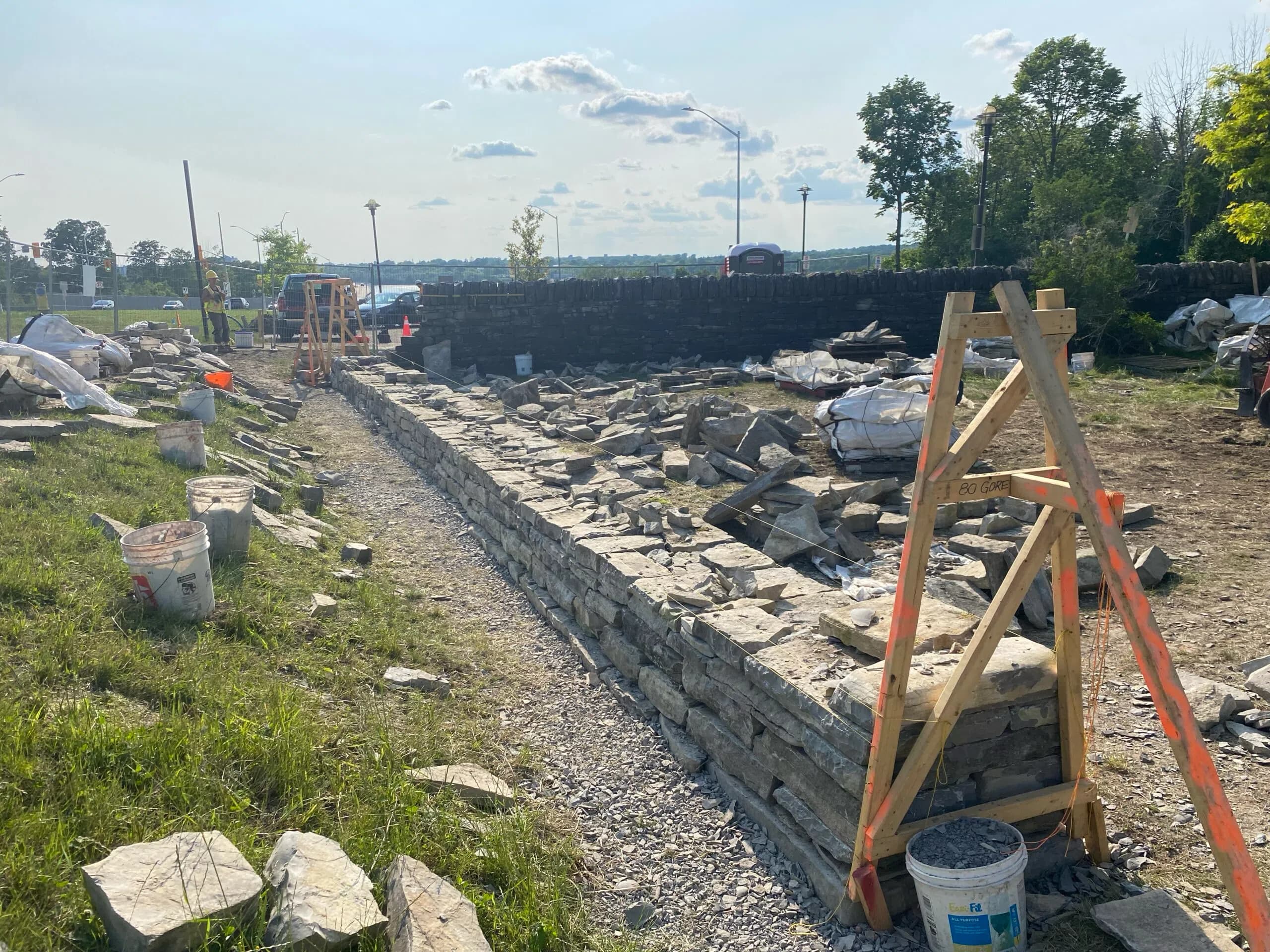 The first courses of the rebuilt wall running away to the west, a timber batter frame standing at the near end and stone laid out on the grass either side.