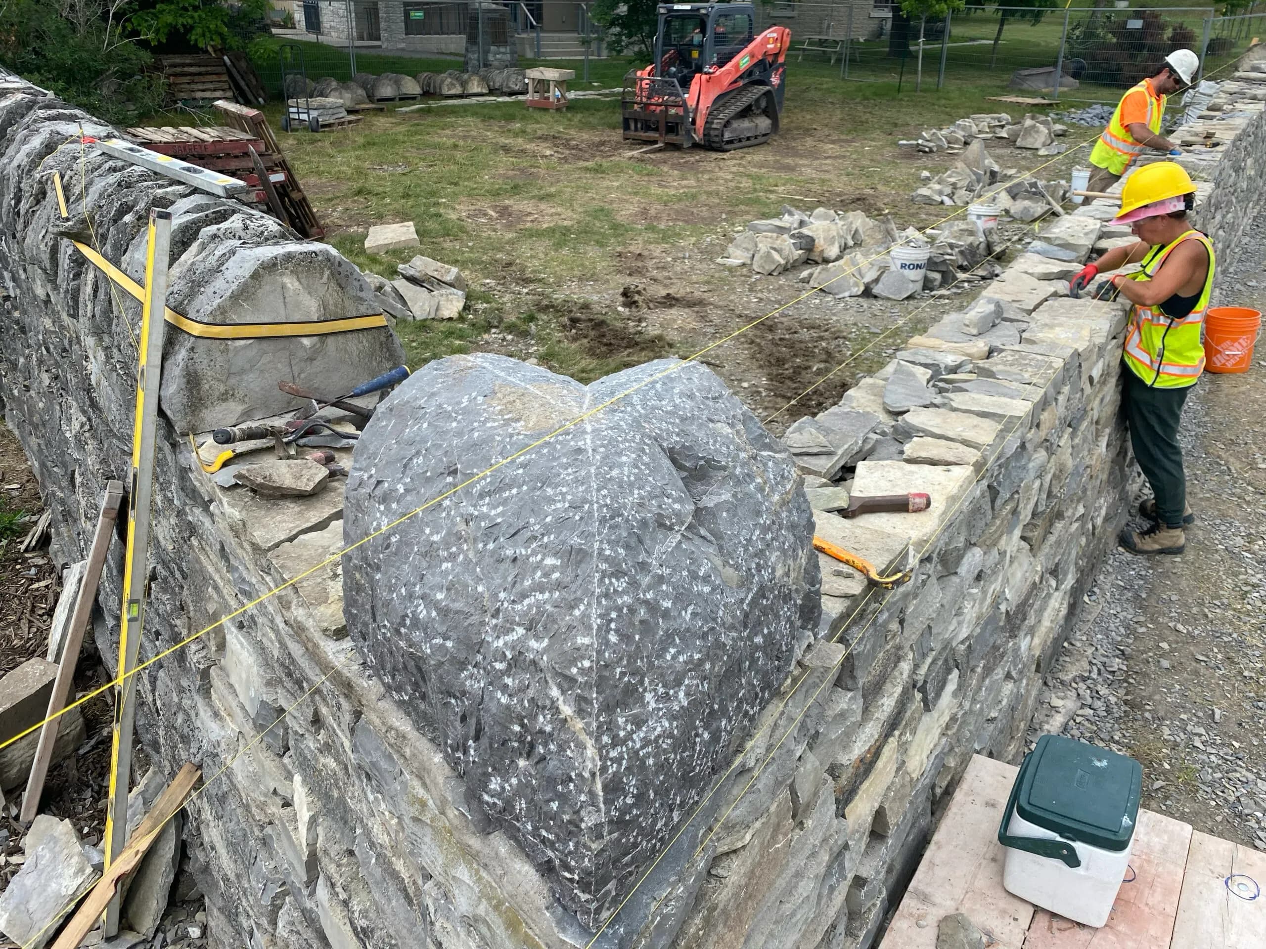 Two wallers setting a large rounded corner stone into the top of the new corner, string lines running along the wall head on both sides of it.