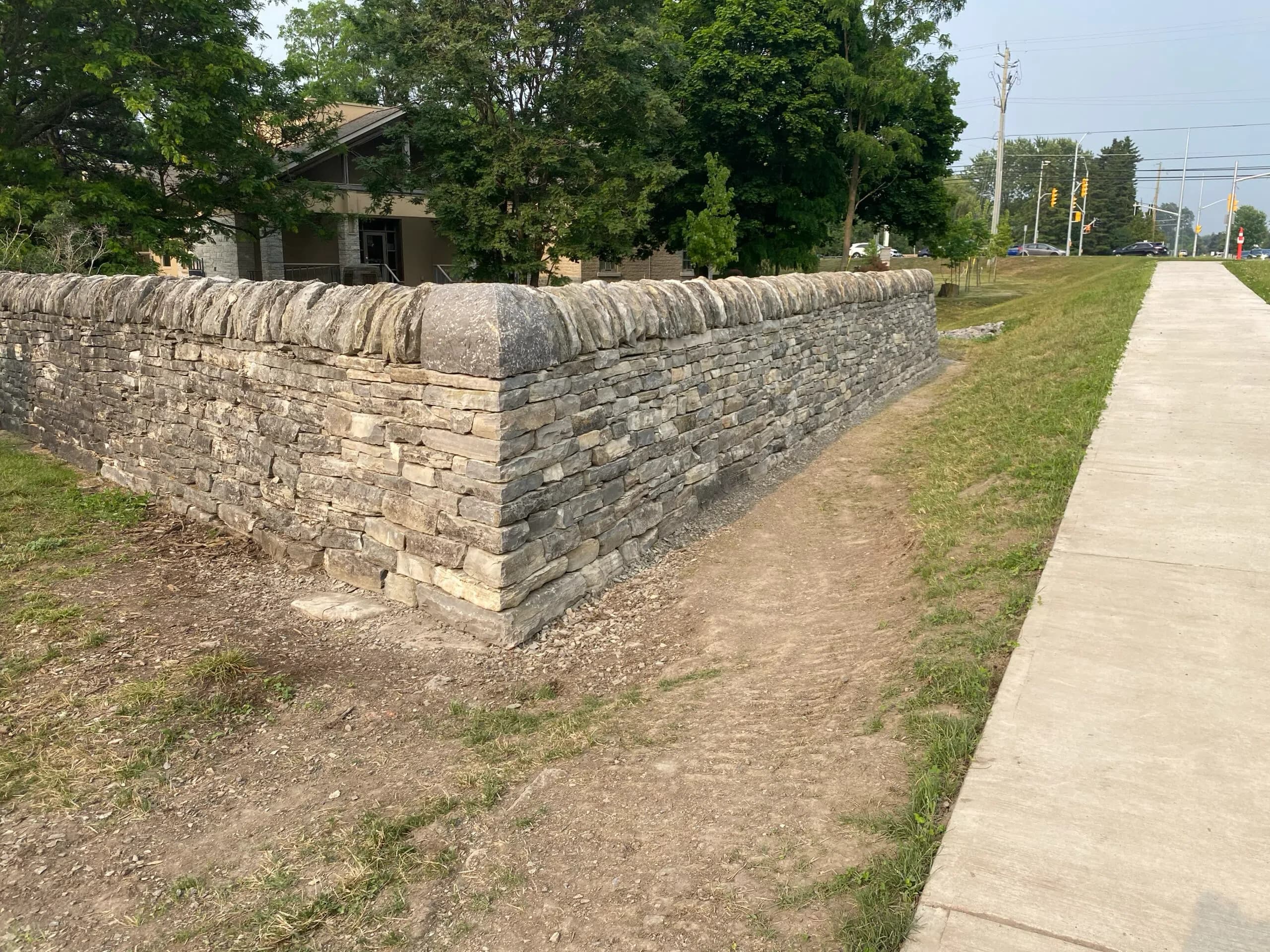 The rebuilt wall meeting its new corner, coping stones running along the top, the c1860 limestone library behind it and the widened road and sidewalk to the right.