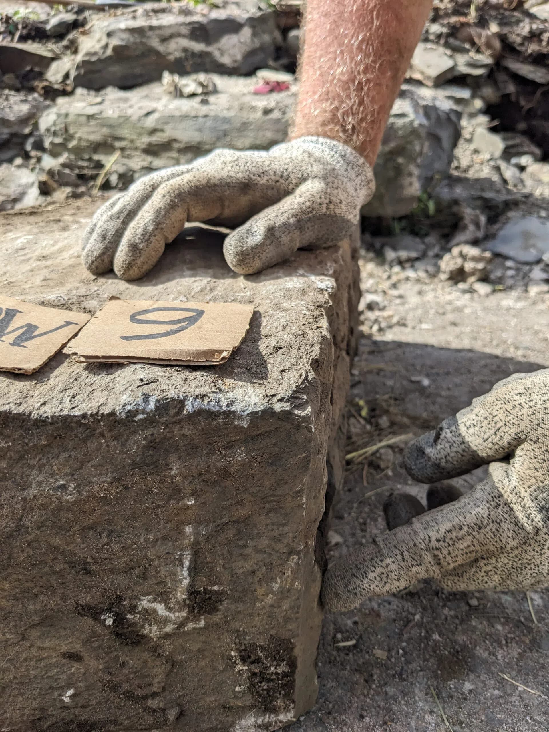Two gloved hands steadying a large slate block on the wall top, a numbered cardboard tag lying on its face.