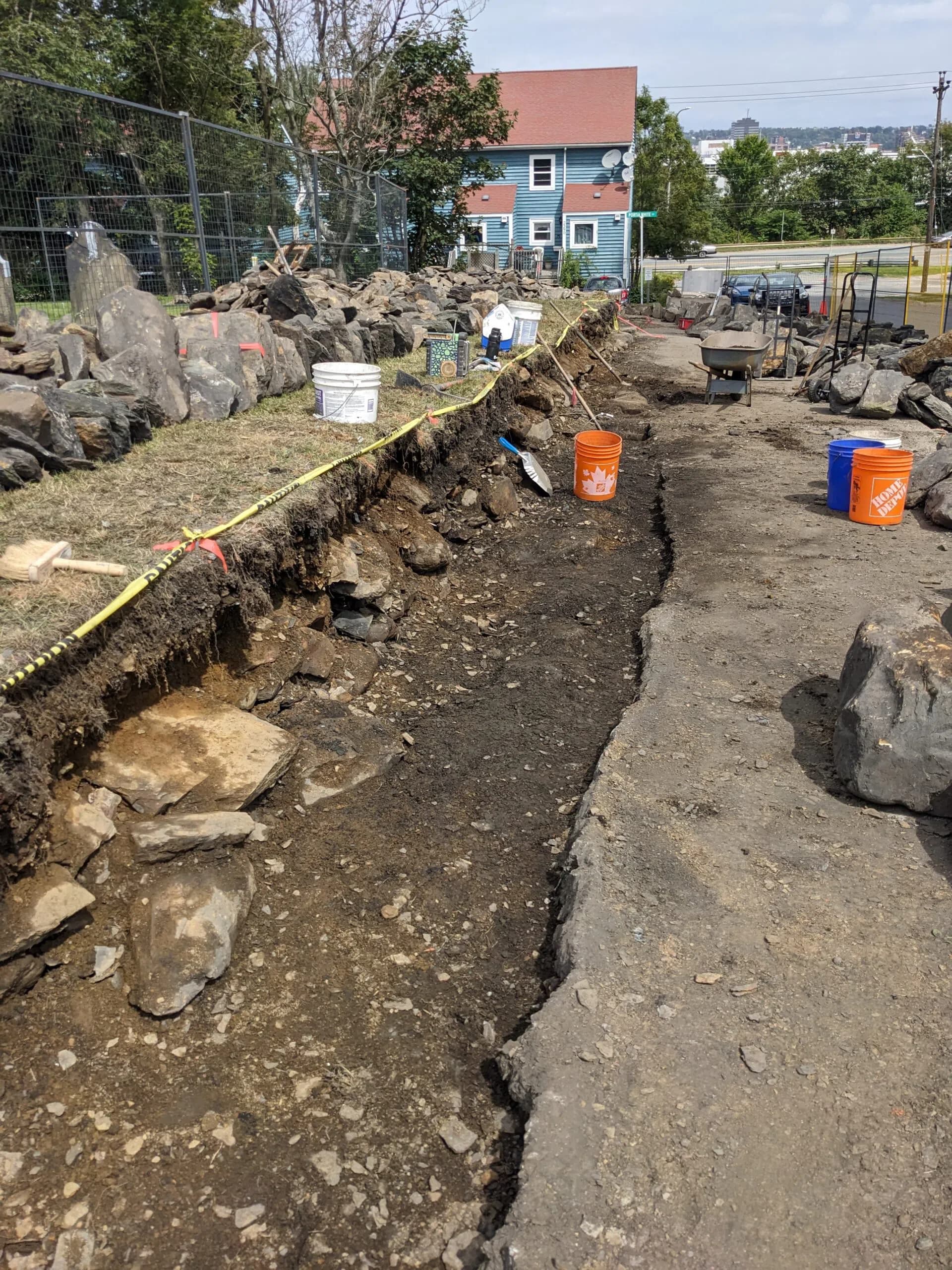 The emptied line of the wall, a long open trench with caution tape strung along it, houses and the harbour beyond.