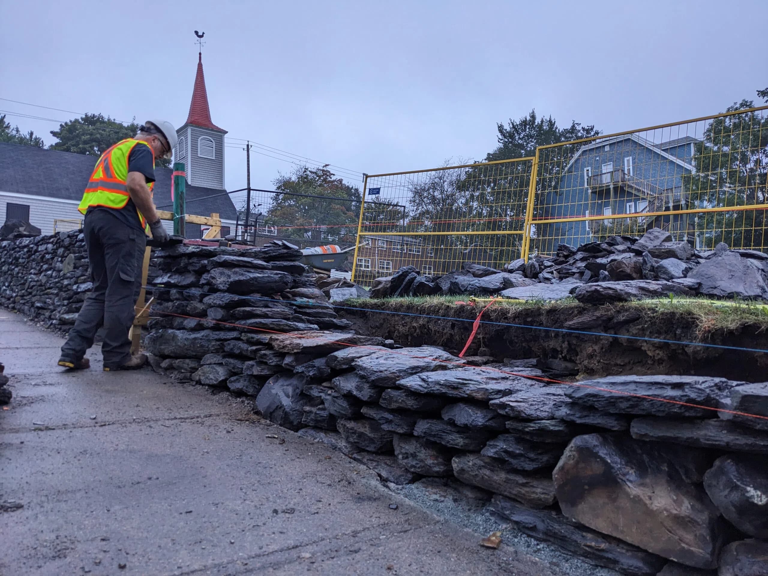 A waller working the top of the part-built wall on a grey wet day, the church and its red steeple beyond and the rebuilt section running away to the right.