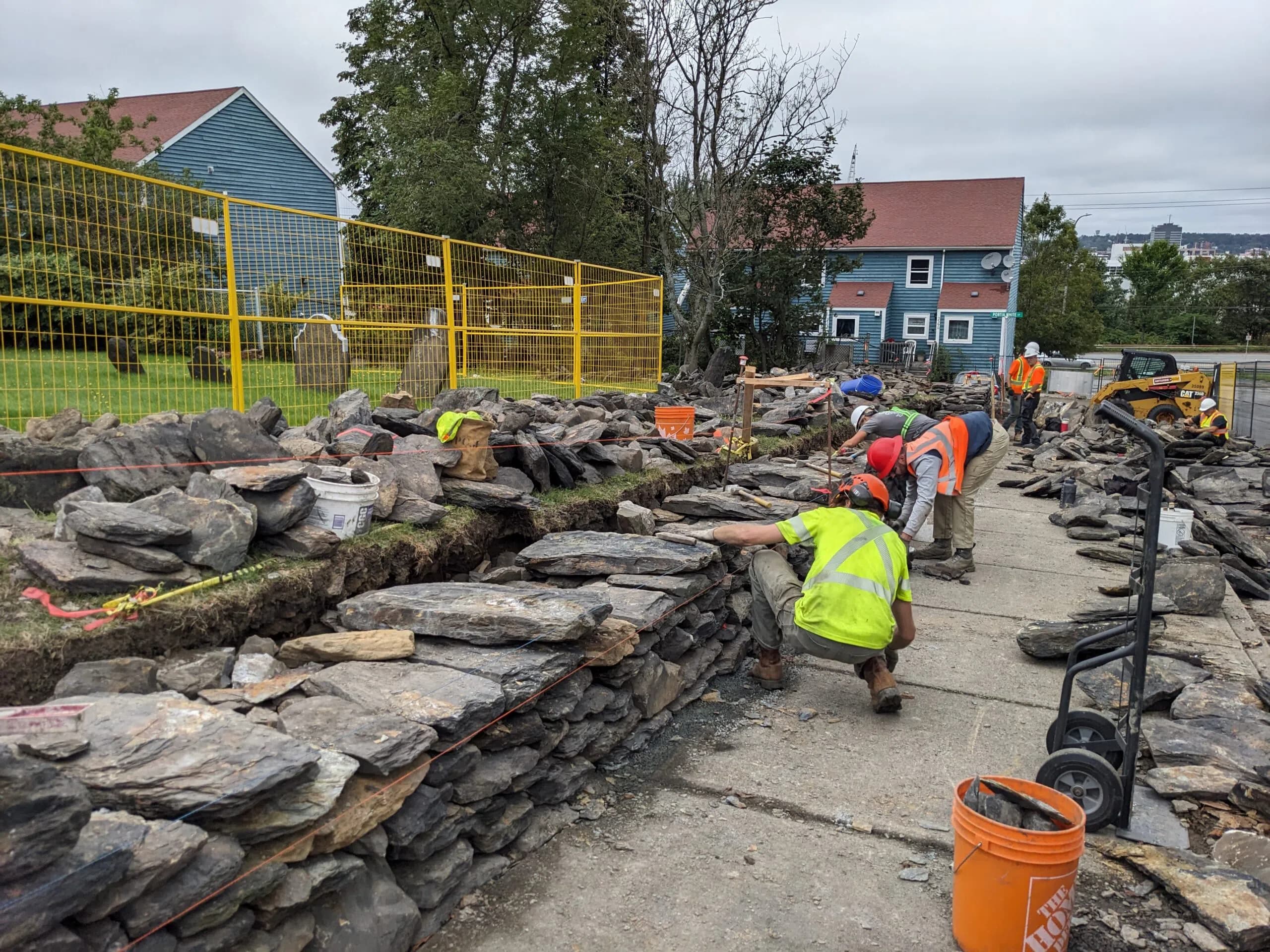 Three of the crew working along the rising wall on the sidewalk, slate stacked either side of them and clapboard houses across the street.