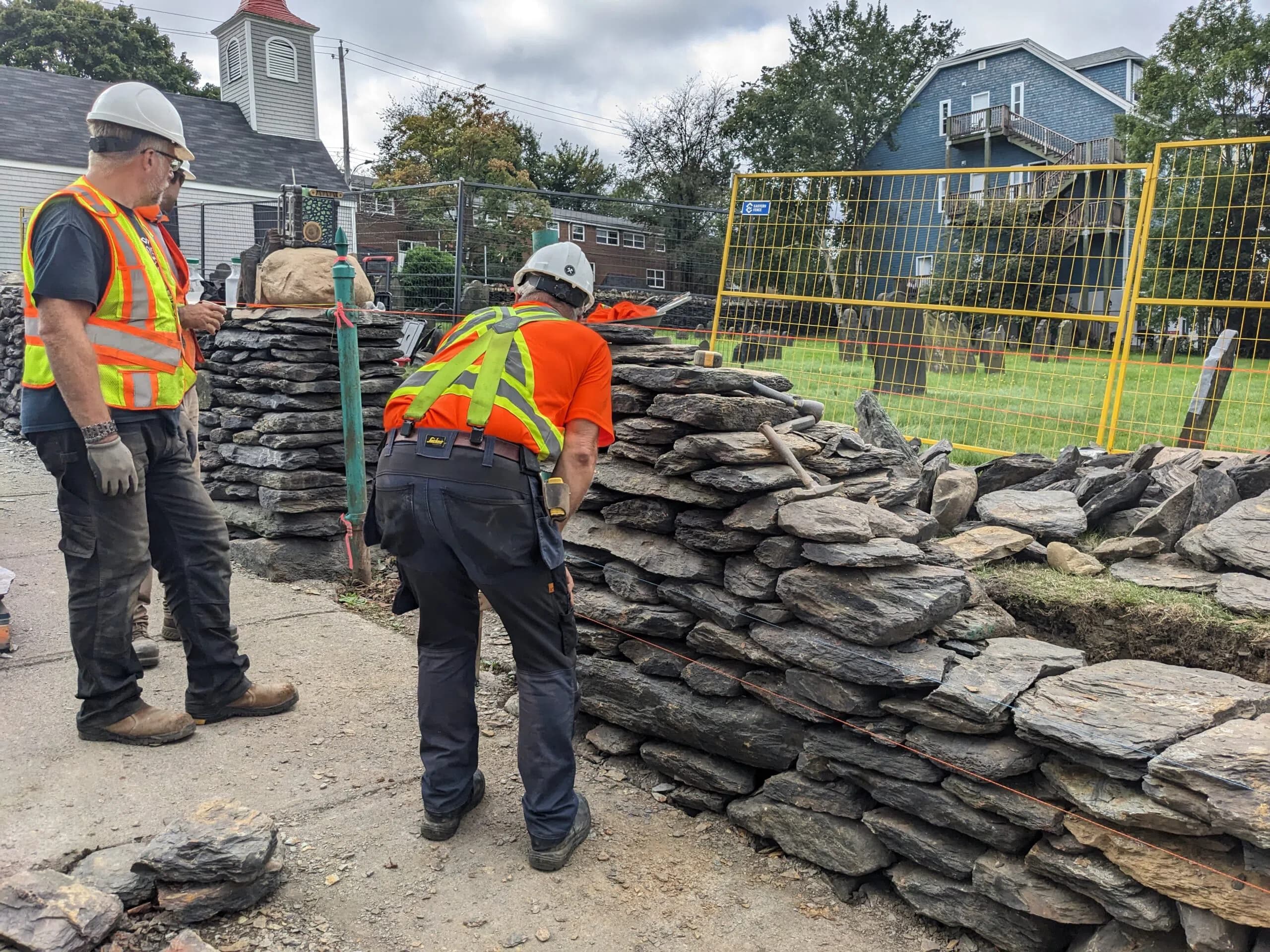 Two wallers at a stack of long slate coping stones beside the finished section of wall, the church and its steeple behind them.