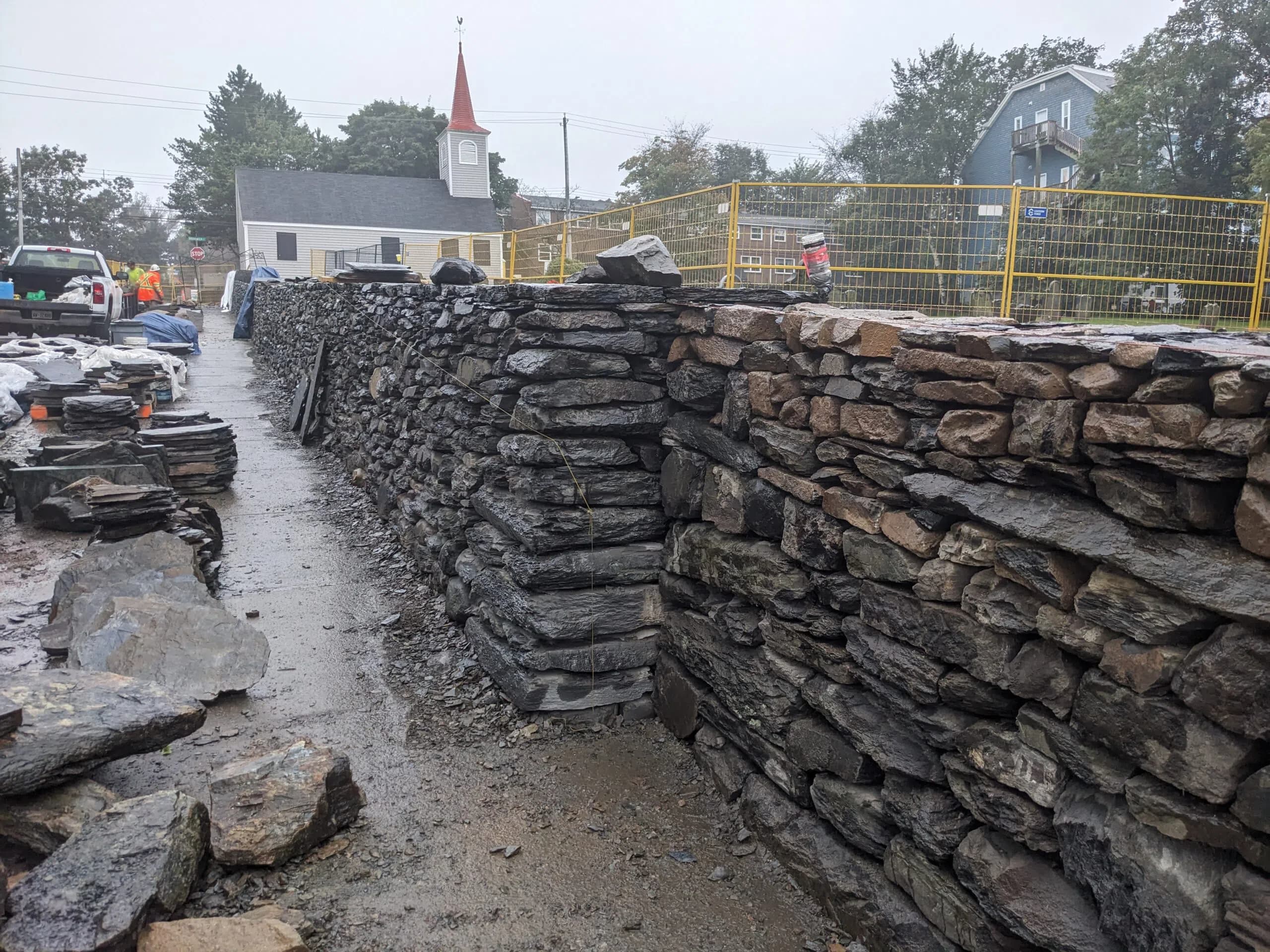 The wall part-built on a wet day, running away up the sidewalk past stacked slate towards the red spire of the Little Dutch Church.