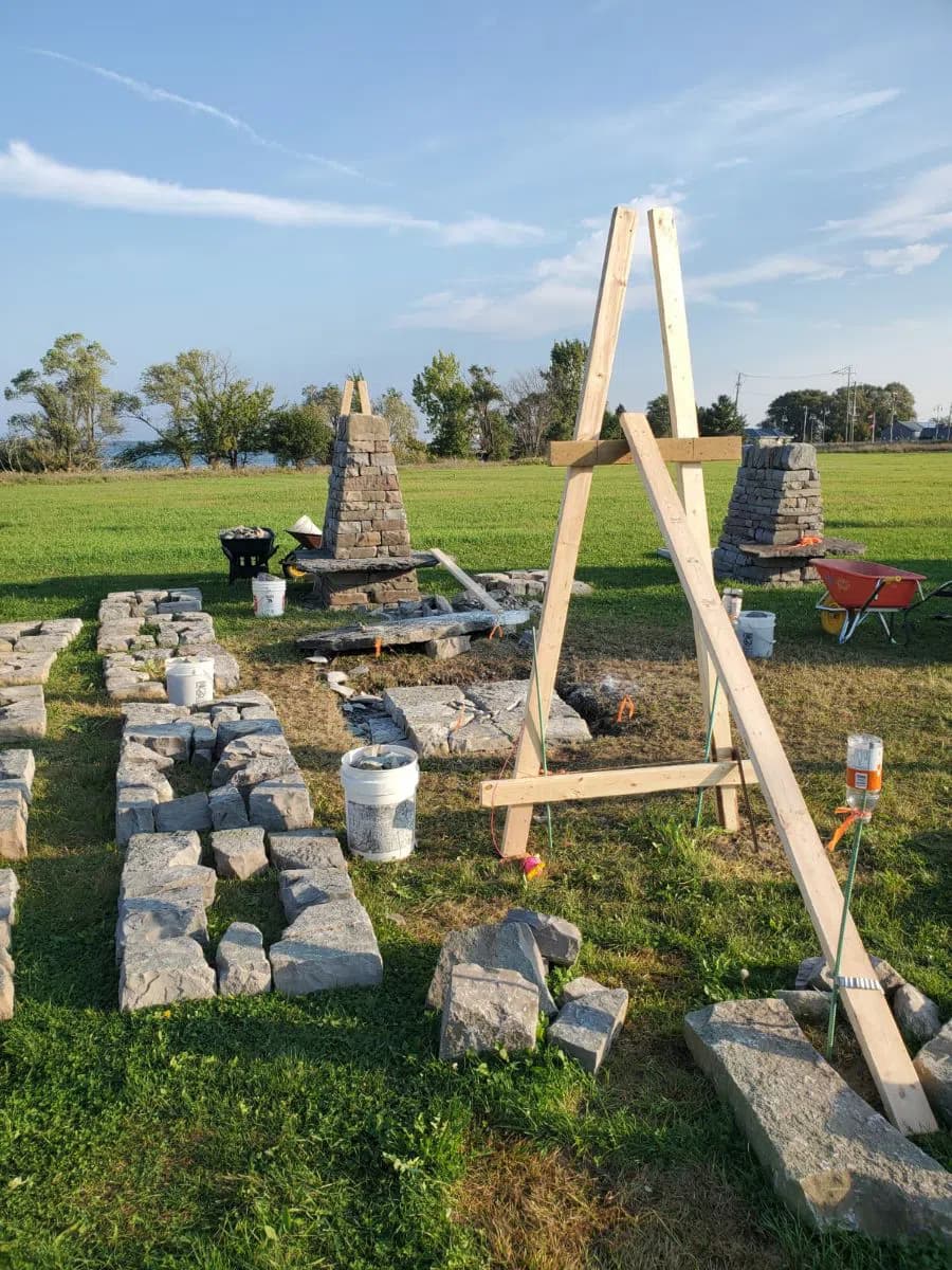 A timber batter frame standing over the cleared pillar footprint in low evening light, the dismantled stone laid out in rows on the grass and two of the standing pillars behind it.
