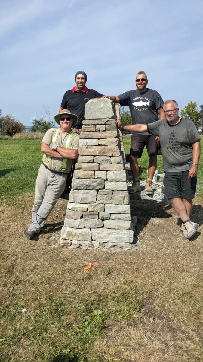 Four wallers standing at the finished pillar, one hand resting on its capstone, the coursing and the taper of the rebuilt structure running down to the grass.