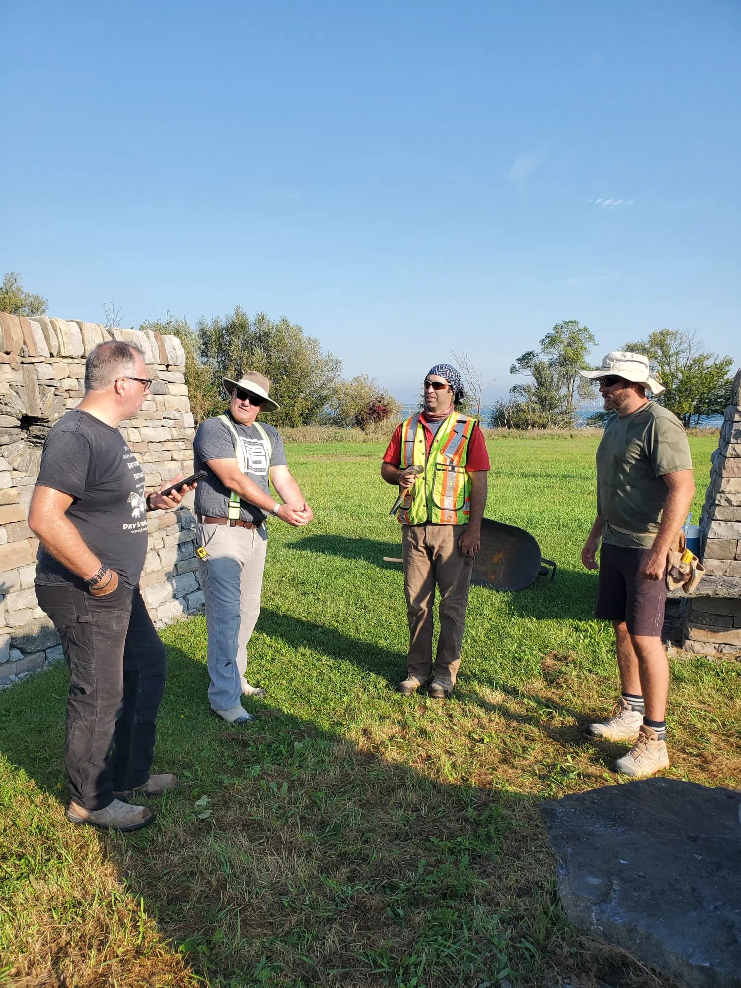 Four of the crew standing and talking on the grass at the solar alignment site, a dry stone wall at the left of the frame, a pillar at the right and the lake behind them.
