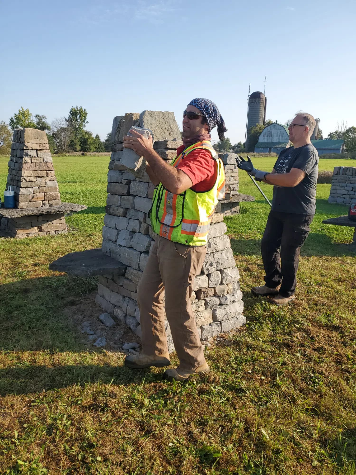 A waller in a hi-vis vest lifting a stone clear of the part-dismantled pillar, a barn and grain silo in the field behind him.