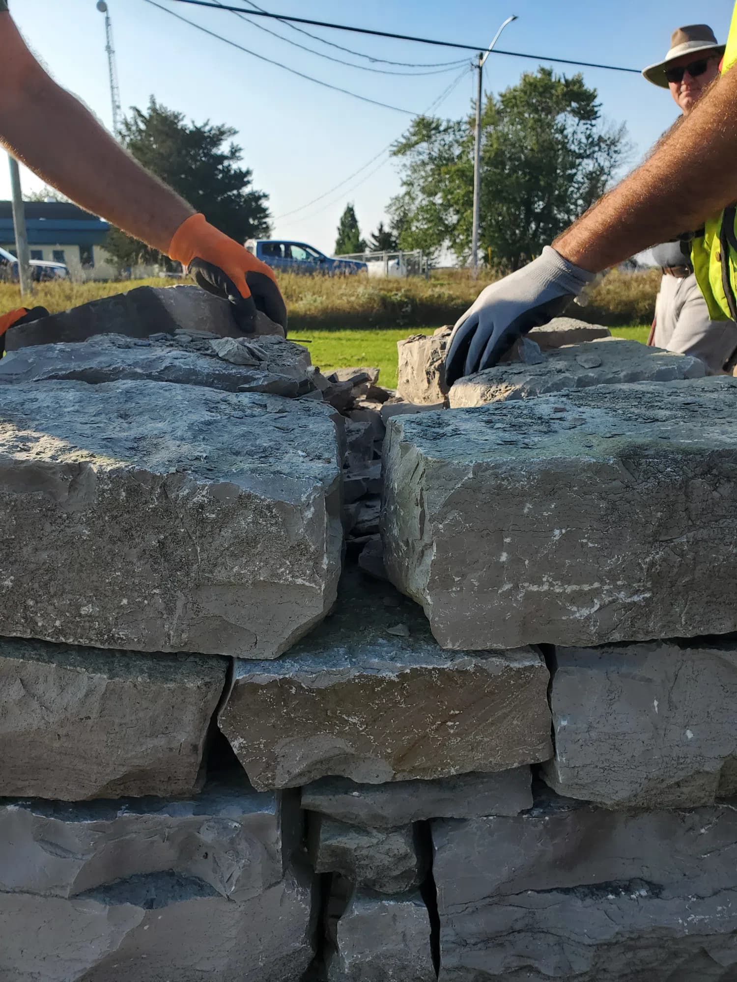 Two large limestone blocks being set side by side on a course, a gloved hand at either end of the frame and the packed hearting visible in the joint between them.
