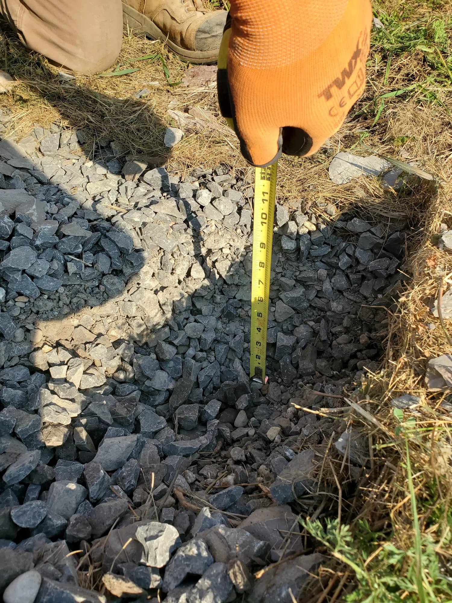 A gloved hand holding a steel tape measure upright in the open excavation, the blade running down past loose stone to the bottom of the hole.