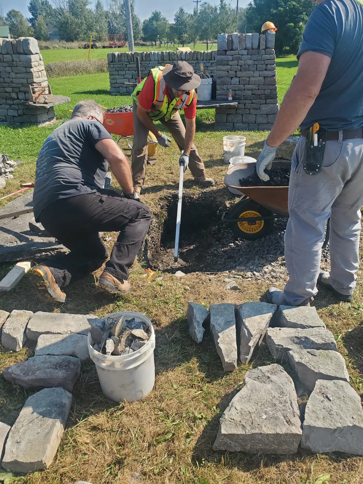Two of the crew digging out the ground beneath the dismantled pillar with a long-handled spade, a wheelbarrow of excavated material beside them.