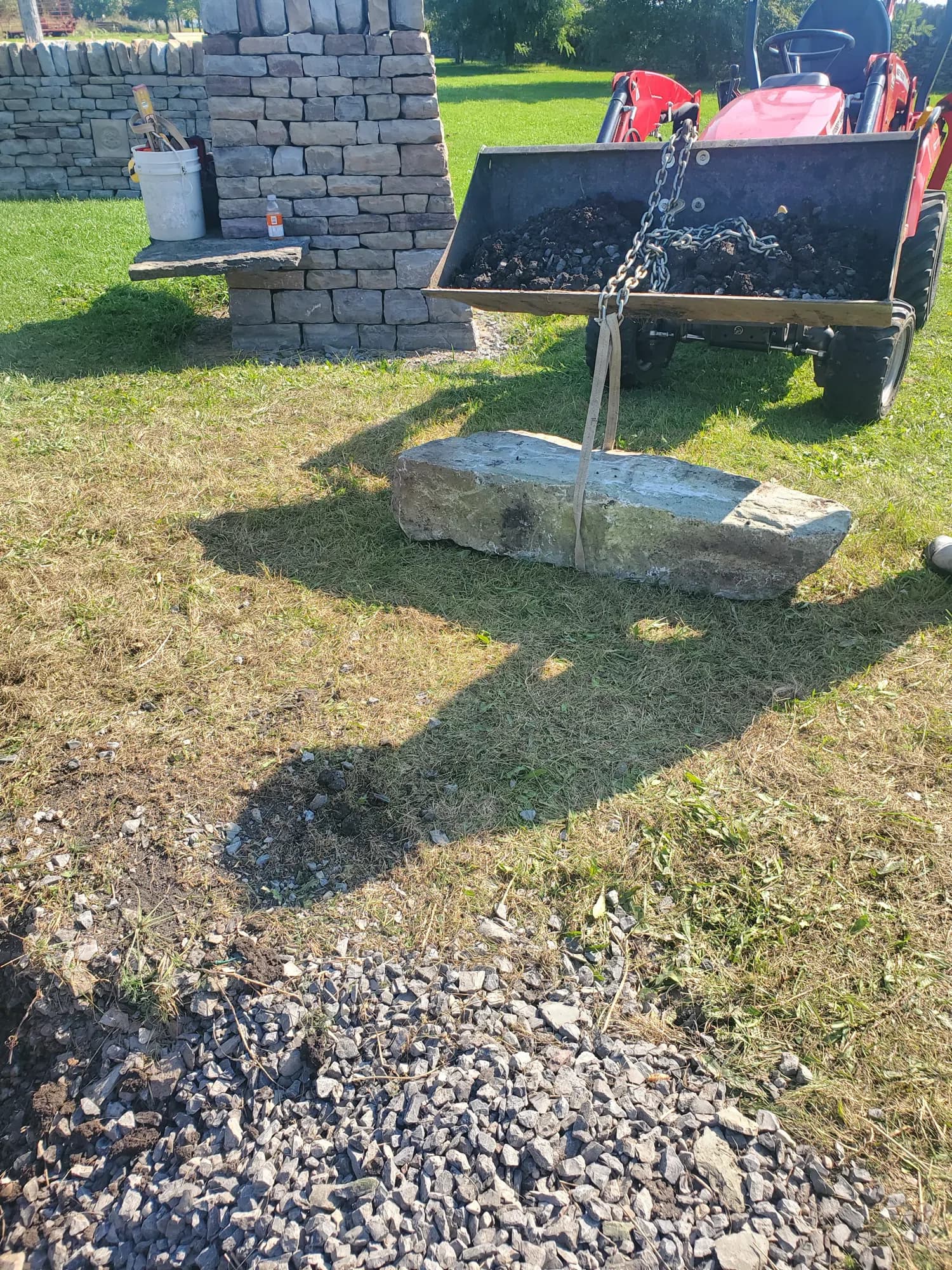 A large flat foundation stone rigged with chains beneath the bucket of a compact tractor, on the grass beside the open excavation.